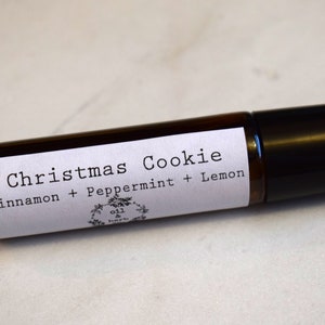 May include: A brown glass roller bottle with a black cap. The label reads "Christmas Cookie Cinnamon + Peppermint + Lemon" with a small logo that says "oil & herbs".