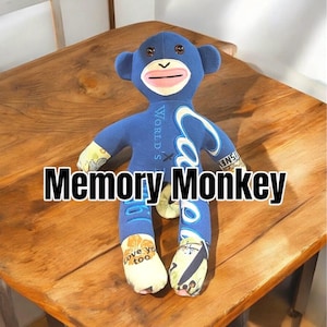 May include: Blue fabric monkey toy with a Coca-Cola logo on its back. The monkey is sitting on a wooden surface. The text "Memory Monkey" is written in black letters below the monkey.
