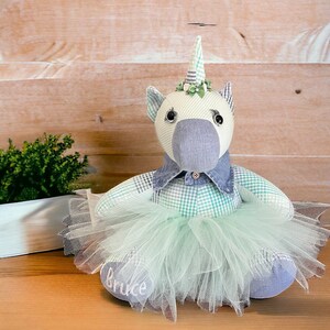 Könnte beinhalten: Ein handgefertigtes Einhorn-Plüschtier mit blau-grünem Karomuster, einem blauen Jeanskragen und einem mintgrünen Tüll-Tutu. Das Einhorn hat ein gestreiftes Horn und ist mit dem Namen "Bruce" auf dem Fuß personalisiert.
