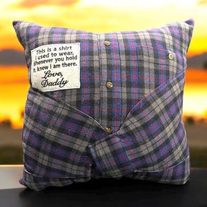 May include: A gray and blue plaid pillow with a white patch sewn on the front. The patch has the text "This is a shirt I used to wear. Whenever you hold it, know I am there. Love, Daddy".