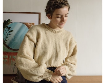 May include: A cream-colored knitted sweater with a round neckline and long sleeves. The sweater has a cropped length and a slightly oversized fit.