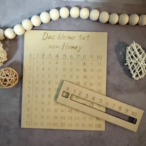 Könnte beinhalten: Eine hölzerne Multiplikationstabelle mit dem Text "Das kleine 1x1 von Henry" wird angezeigt. Ein hölzernes Lineal liegt auf dem Tisch. Der Tisch hat Zahlen von 1 bis 10. Eine Kette aus Holzperlen und dekorativen Objekten befindet sich im Hintergrund.
