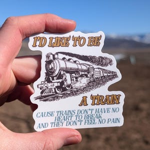 May include: A sticker featuring a vintage train illustration and the text "I'D LIKE TO BE A TRAIN CAUSE TRAINS DON'T HAVE NO HEART TO BREAK AND THEY DON'T FEEL NO PAIN." The sticker is held up against a blue sky and a blurred landscape.