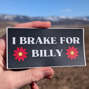 May include: A black rectangular sticker with white text that reads "I BRAKE FOR BILLY". The sticker has two red and yellow flower graphics. The sticker is held in a hand against a backdrop of a blue sky and mountains.