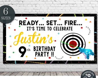 May include: A white and black birthday banner with colourful geometric shapes and a target with a toy gun. The banner says "Ready... Set... Fire... It's time to celebrate Justin's 9th Birthday Party!!"