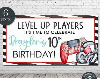 May include: A black and white striped banner with red and black stripes at the top and bottom. The banner says "Level Up Players It's Time To Celebrate Brayden's 10th Birthday!" in black text with a blue "Brayden's" and a black "10th". There is a red and black video game controller and a pair of black headphones with red accents on the banner.