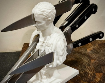 May include: A white bust sculpture with multiple knives inserted through it. The knives have silver blades and black handles. The sculpture is on a white base, and the background is a wooden surface.