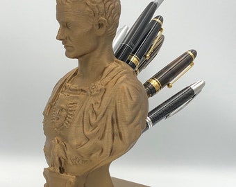 Puede incluir: Un busto de color bronce de un emperador romano, que sirve como portalápices. Varios bolígrafos de varios diseños y colores están insertados en el busto. El busto está montado sobre una base cuadrada, creando un accesorio de escritorio funcional y decorativo.