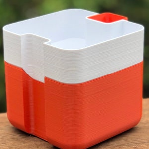 May include: A two-tone container with a square shape. The bottom half is orange, and the top half is white. A small orange rectangular compartment is attached to the top rim. The container is sitting on a wooden surface.