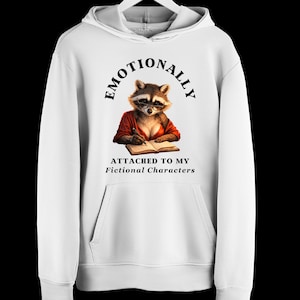 Puede incluir: Una sudadera con capucha blanca que presenta un gráfico de un mapache con gafas, escribiendo en un libro y vistiendo una prenda roja. El texto EMOTIONALLY ATTACHED TO MY Fictional Characters aparece en letras negras sobre y bajo la ilustración.