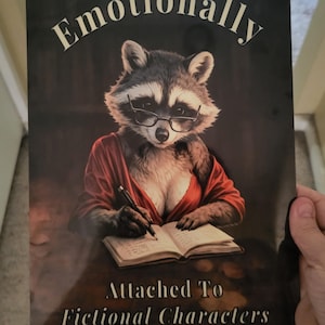 May include: A rectangular metal sign featuring a raccoon wearing glasses and a red shirt, writing in an open book. The text at the top reads Emotionally and the text at the bottom reads Attached To Fictional Characters in a serif font.