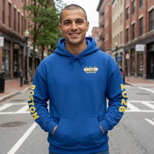 Puede incluir: Una sudadera con capucha azul con bolsillo delantero y capucha con cordón. La sudadera tiene la palabra "BOSTON" impresa en amarillo en la manga izquierda y el año "2026" en la manga derecha. La parte delantera de la sudadera tiene la palabra "BOSTON" y la fecha "APRIL 26, 2026" impresas en amarillo.