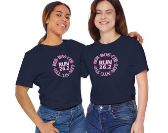 May include: Two women wearing navy blue t-shirts with a pink circular graphic that reads "RUN 26.2" and the cities BER, BOS, CHI, LON, NYC, and TYO. The women are smiling and wearing blue jeans.