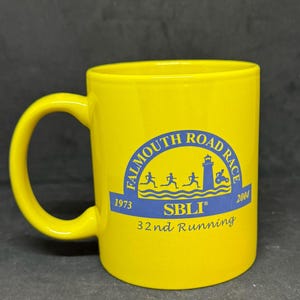 May include: A yellow ceramic mug with a curved handle. The mug features a blue circular design with the text "Falmouth Road Race" and the years "1973" and "2004". The text "SBLI" and "32nd Running" are also visible.