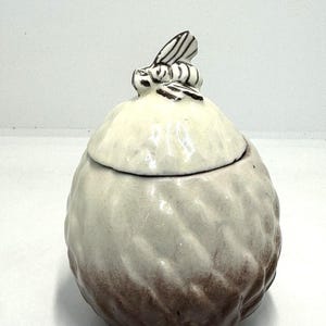 May include: A ceramic honey pot with a lid, featuring a bee figurine on top. The pot is a light beige color, with a textured surface and a darker brown base. The bee is black and white.