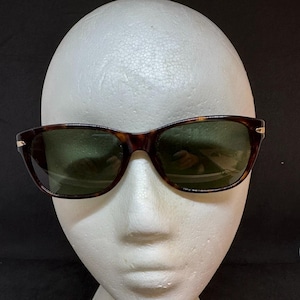 May include: Tortoiseshell sunglasses with green lenses are displayed on a white mannequin head. The sunglasses have a classic rectangular frame with a subtle gold accent on the temples. The lenses are a dark green color.