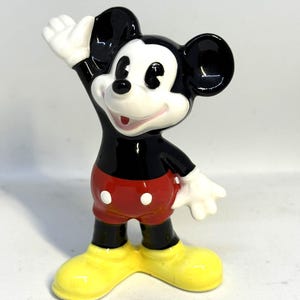May include: A ceramic figurine of Mickey Mouse, a classic Disney character. Mickey is depicted with black ears, a black torso, red shorts, yellow shoes, and white gloves. He is waving with his right hand.