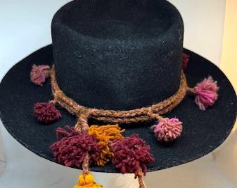 Pode incluir: Um chapéu de lã preto com aba larga e copa cilíndrica. O chapéu é adornado com uma faixa trançada marrom e borlas coloridas em tons de rosa, amarelo e bordô, adicionando um toque decorativo.
