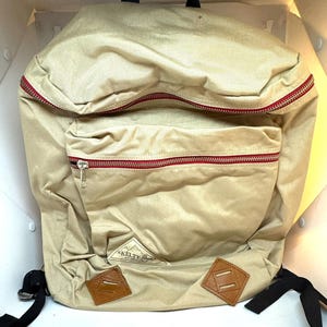 May include: Tan backpack with red zippers and a front pocket. Features brown leather accents and the "KELTY Pack" logo. Includes black shoulder straps and a top handle.