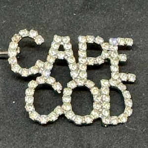 May include: A silver-toned pin featuring the words "CAPE COD" spelled out in sparkling, clear crystals. The pin has a long, thin needle for attachment. The design is a decorative accessory.