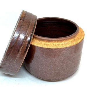 May include: A brown ceramic jar with a lid, set against a white background. The jar has a cylindrical shape with a lighter-colored band around the rim. The lid is partially off, revealing the interior. The jar's exterior has a speckled texture.