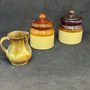 May include: A ceramic creamer pitcher with a mottled yellow and brown glaze alongside two cylindrical storage jars with lids. One jar features a tan base with a brown glazed top, while the other has a tan base with a dark brown glazed lid.