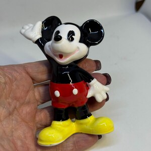 May include: A ceramic Mickey Mouse figurine. The figure has black ears, a black body, red shorts with white buttons, white gloves, and yellow shoes. Mickey is waving with a smile.