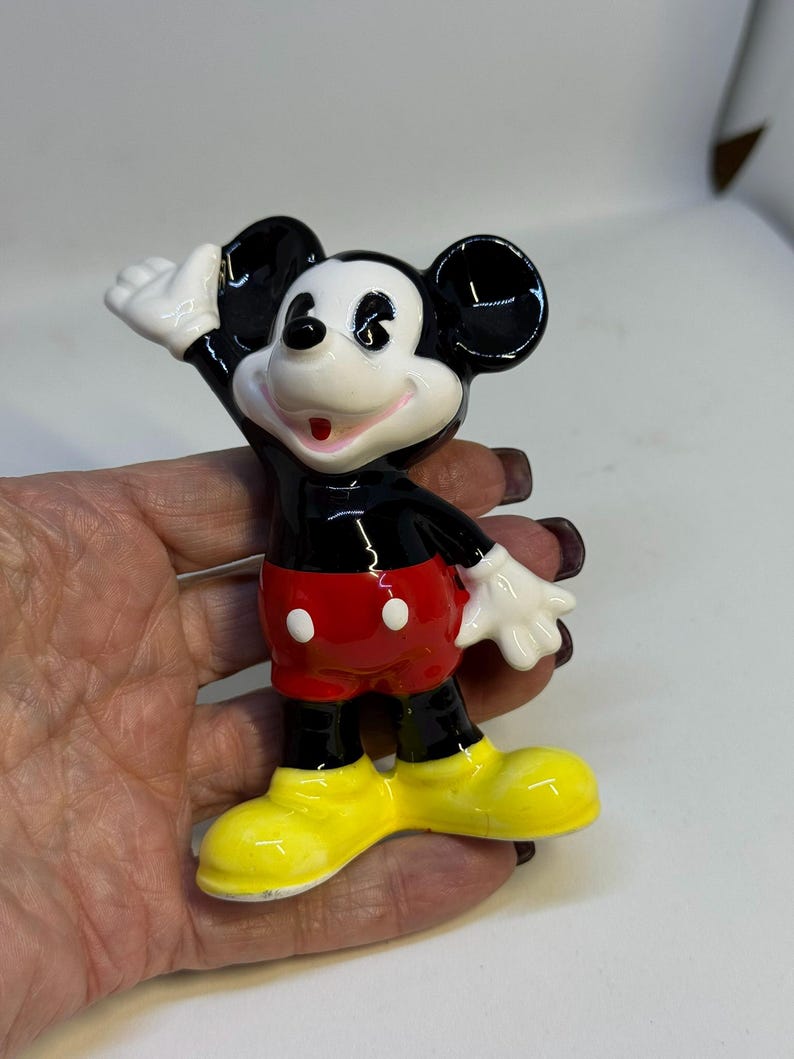 May include: A ceramic Mickey Mouse figurine. The figure has black ears, a black body, red shorts with white buttons, white gloves, and yellow shoes. Mickey is waving with a smile.