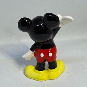 May include: A ceramic figurine of Mickey Mouse, viewed from the back. Mickey is wearing red shorts with white buttons, yellow shoes, and white gloves. His black ears and head are prominent, with one arm raised.