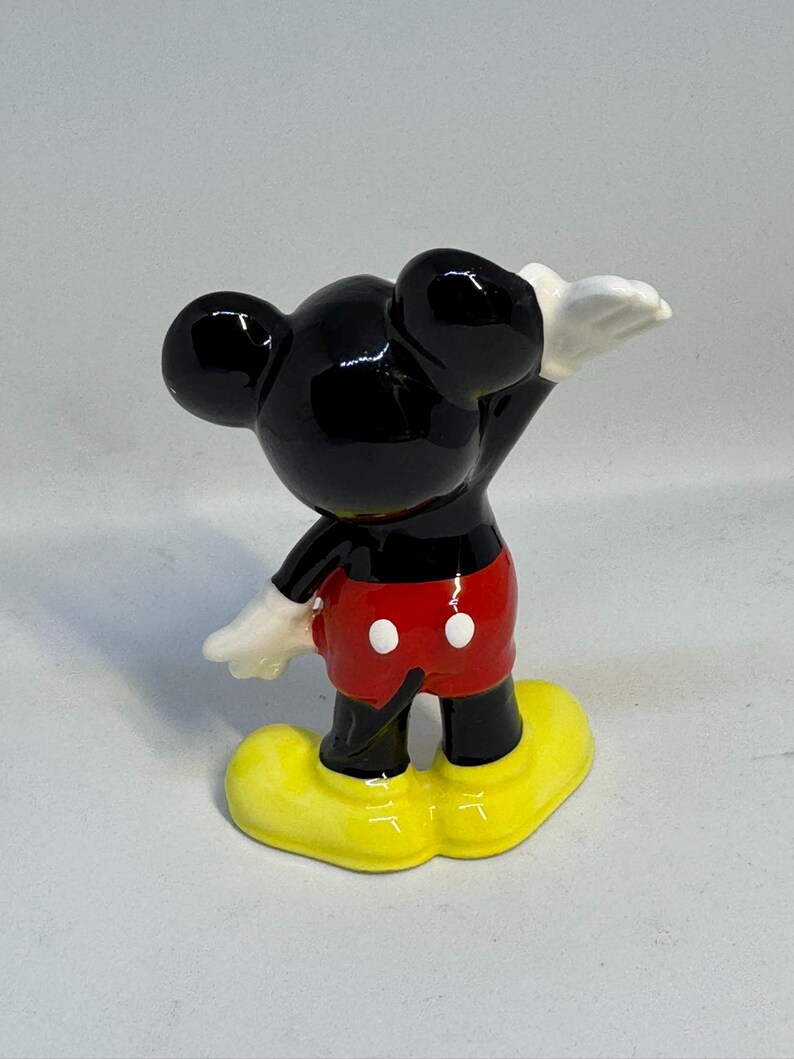 May include: A ceramic figurine of Mickey Mouse, viewed from the back. Mickey is wearing red shorts with white buttons, yellow shoes, and white gloves. His black ears and head are prominent, with one arm raised.