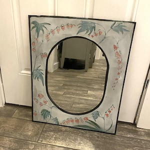 May include: Oval mirror with a black frame and a floral design. The mirror's frame is painted light blue with green leaves and red flowers. The mirror is resting on a wooden floor.