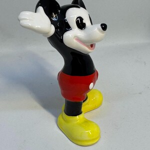 May include: A ceramic Mickey Mouse figurine. The figure has a black head and body, red shorts, yellow shoes, and white gloves. The figurine is posed with its arms up and mouth open, and is standing on a white surface.
