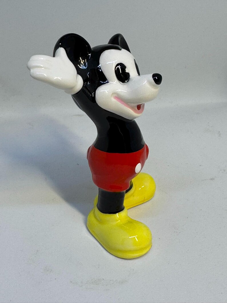 May include: A ceramic Mickey Mouse figurine. The figure has a black head and body, red shorts, yellow shoes, and white gloves. The figurine is posed with its arms up and mouth open, and is standing on a white surface.