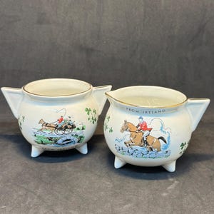 May include: Two cream-colored ceramic creamers with gold trim and small feet. One creamer features a horse-drawn carriage scene, the other a rider on a horse with the text "FROM IRELAND". Both have shamrock designs.