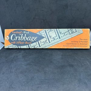 May include: A rectangular box for Cardinal's Wood Cribbage with a 2-player board. The orange and black packaging features the text 'Cardinal's Wood Cribbage with 2-Player Board' and 'The most exciting 2-Handed Card Game in America! Complete with Pegs and Instructions!'