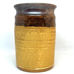 May include: A handmade ceramic utensil holder. The top section is a dark brown, transitioning to a lighter tan color on the lower portion. The cylindrical shape has horizontal grooves, and the rim is slightly flared.