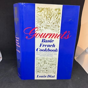 May include: A hardcover cookbook titled Gourmet's Basic French Cookbook by Louis Diat. The cover features a central white vertical panel with red and blue text, flanked by solid blue sides. Decorative line art illustrations of food appear above and below the title.