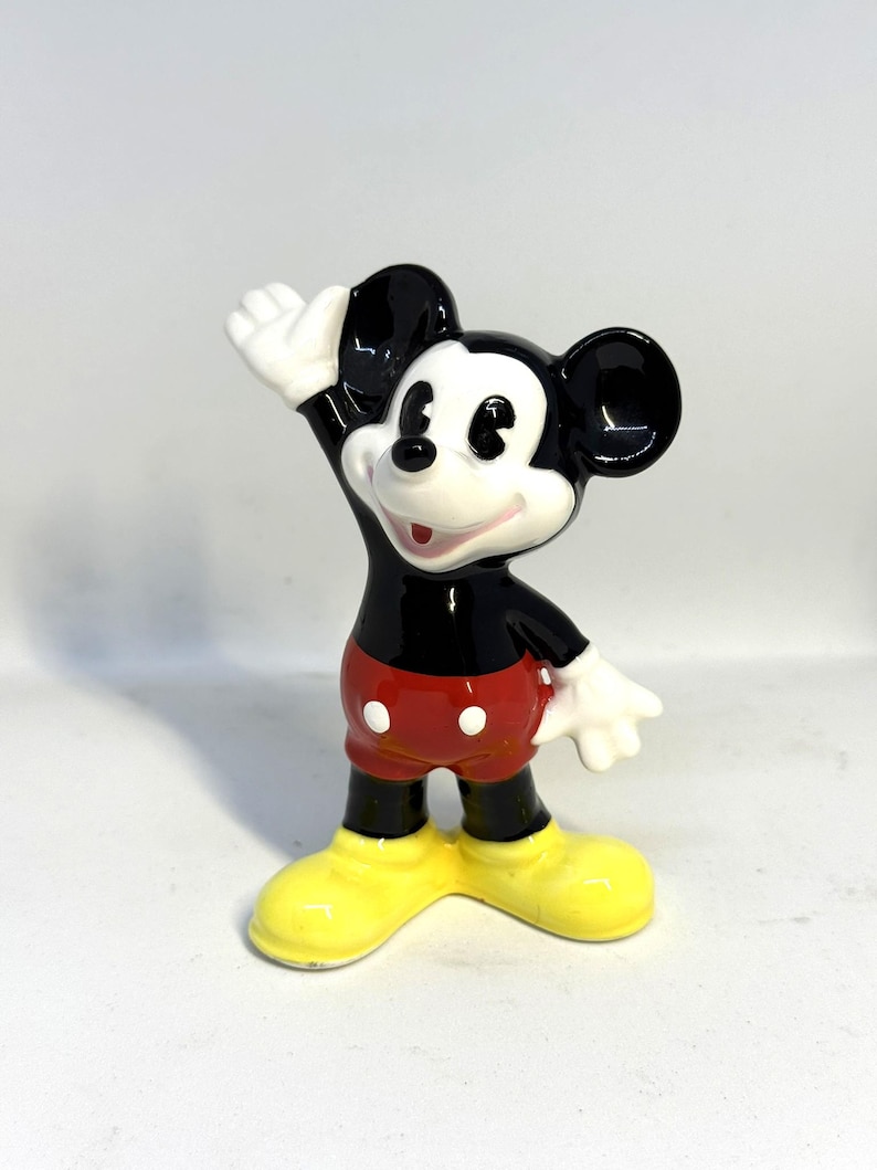 May include: A ceramic figurine of Mickey Mouse, a classic Disney character. Mickey is depicted with black ears, a black torso, red shorts, yellow shoes, and white gloves. He is waving with his right hand.