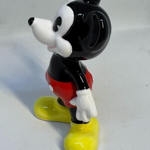 May include: A ceramic figurine of Mickey Mouse, a classic cartoon character. The figure is in a standing pose, with a black head and ears, white face, and red shorts. The shoes are bright yellow, and the gloves are white.