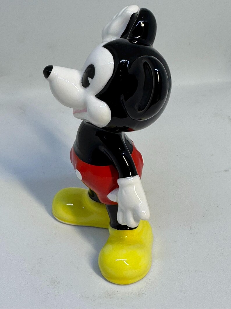 May include: A ceramic figurine of Mickey Mouse, a classic cartoon character. The figure is in a standing pose, with a black head and ears, white face, and red shorts. The shoes are bright yellow, and the gloves are white.