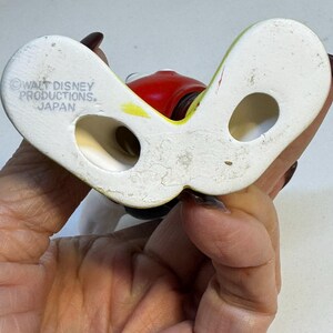 May include: A vintage ceramic figurine base, featuring a white, curved design with two holes. The figurine has a red head and yellow accents. The base is marked "WALT DISNEY PRODUCTIONS. JAPAN".