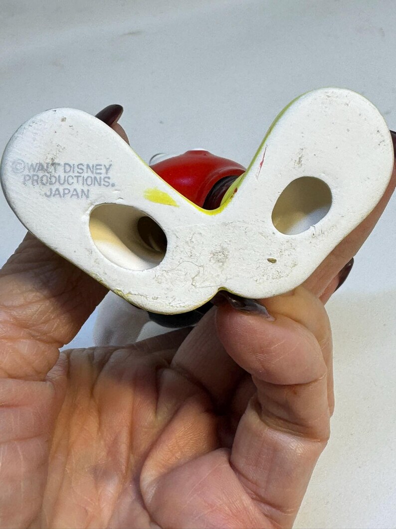 May include: A vintage ceramic figurine base, featuring a white, curved design with two holes. The figurine has a red head and yellow accents. The base is marked "WALT DISNEY PRODUCTIONS. JAPAN".