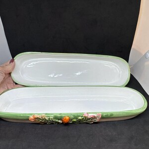 May include: A long, narrow, oval ceramic serving dish with a white interior and a light green rim. The exterior features a decorative relief of green leaves, pink flowers, and a small orange fruit centered on the side of the dish.