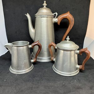 May include: A three-piece metal tea set consisting of a tall coffee pot, a creamer, and a lidded sugar bowl. Each piece features a brushed silver-toned metal body and a curved, dark brown wooden handle. The coffee pot has a matching metal lid.