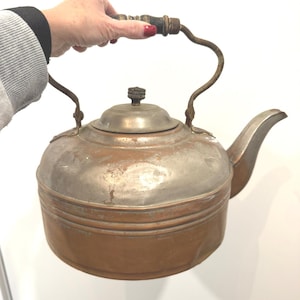 May include: Antique copper and metal kettle with a curved handle and spout. The kettle has a wooden handle and a lid with a decorative knob. The metal has a weathered appearance, showing signs of age and use. A vintage kitchenware item.