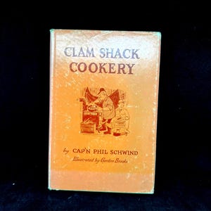 May include: A hardcover book titled Clam Shack Cookery by Cap'n Phil Schwind, illustrated by Gordon Brooks. The orange cover features a dark brown line drawing of a person cooking at a stove, with the title printed in bold dark brown lettering.
