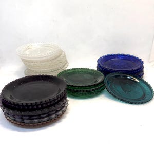 May include: A collection of vintage glass plates in various colors, including clear, black, green, blue, and teal. Each plate has a scalloped edge and a decorative embossed design. The plates are stacked in multiple piles.