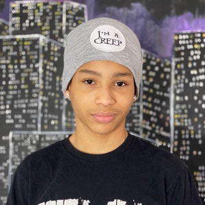 May include: A young person wearing a gray beanie with a black patch that says "I'm a Creep" and a black t-shirt with the word "FELON" printed in white.