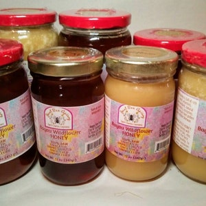 May include: A collection of glass jars filled with honey, each sealed with a red lid. The jars vary in size and honey color, ranging from light amber to dark brown. Labels read "Bayou Wildflower Honey."