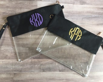 Puede incluir: Dos estuches negros con frentes transparentes y correas de muñeca. Uno tiene un monograma morado, el otro un monograma dorado. Ambos tienen cremalleras doradas y correas de muñeca negras. Los estuches son rectangulares.